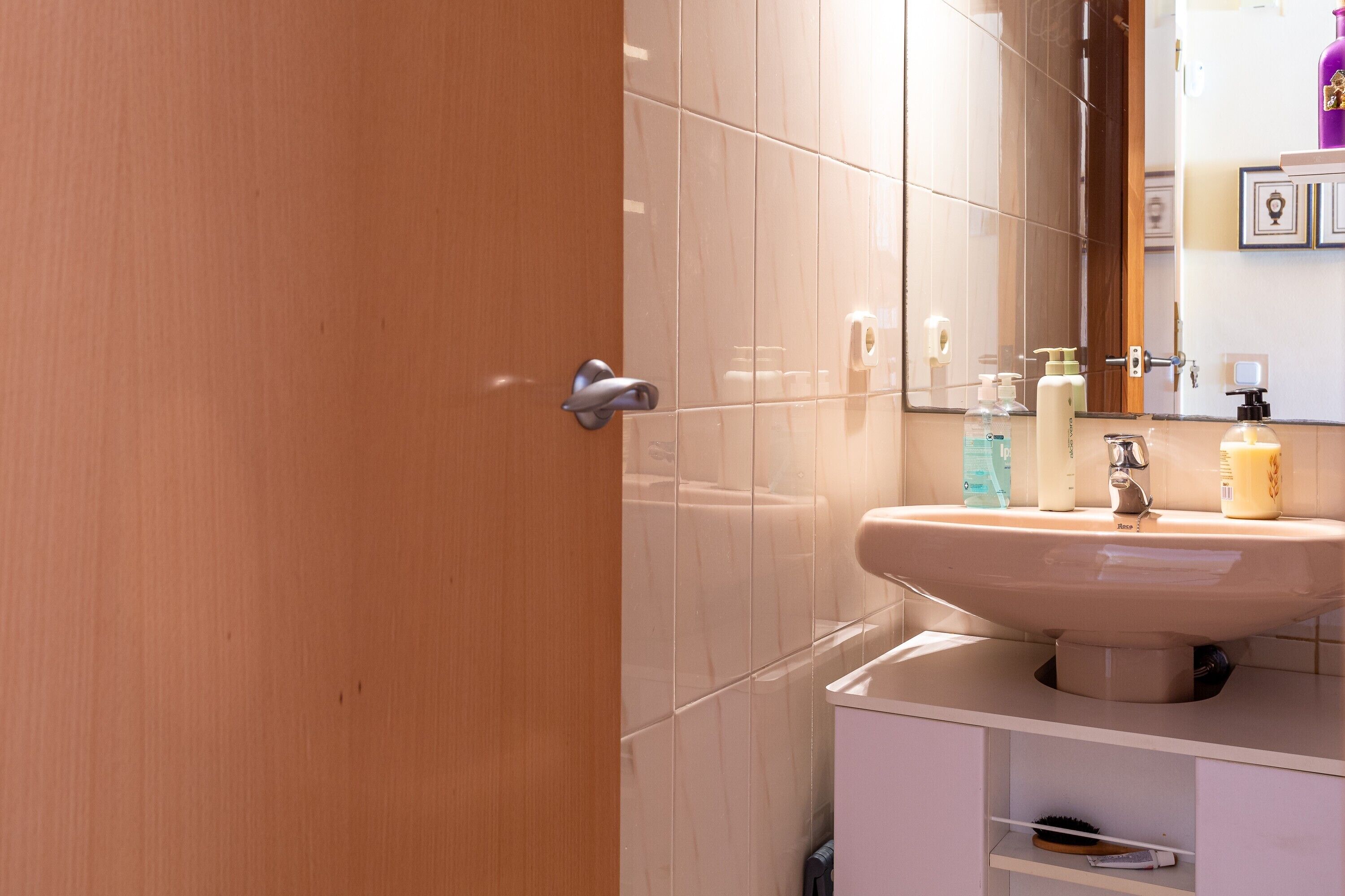 Ein Badezimmer mit Holztür, Waschbecken mit Spiegel und Toilettenartikeln auf der Ablagefläche.