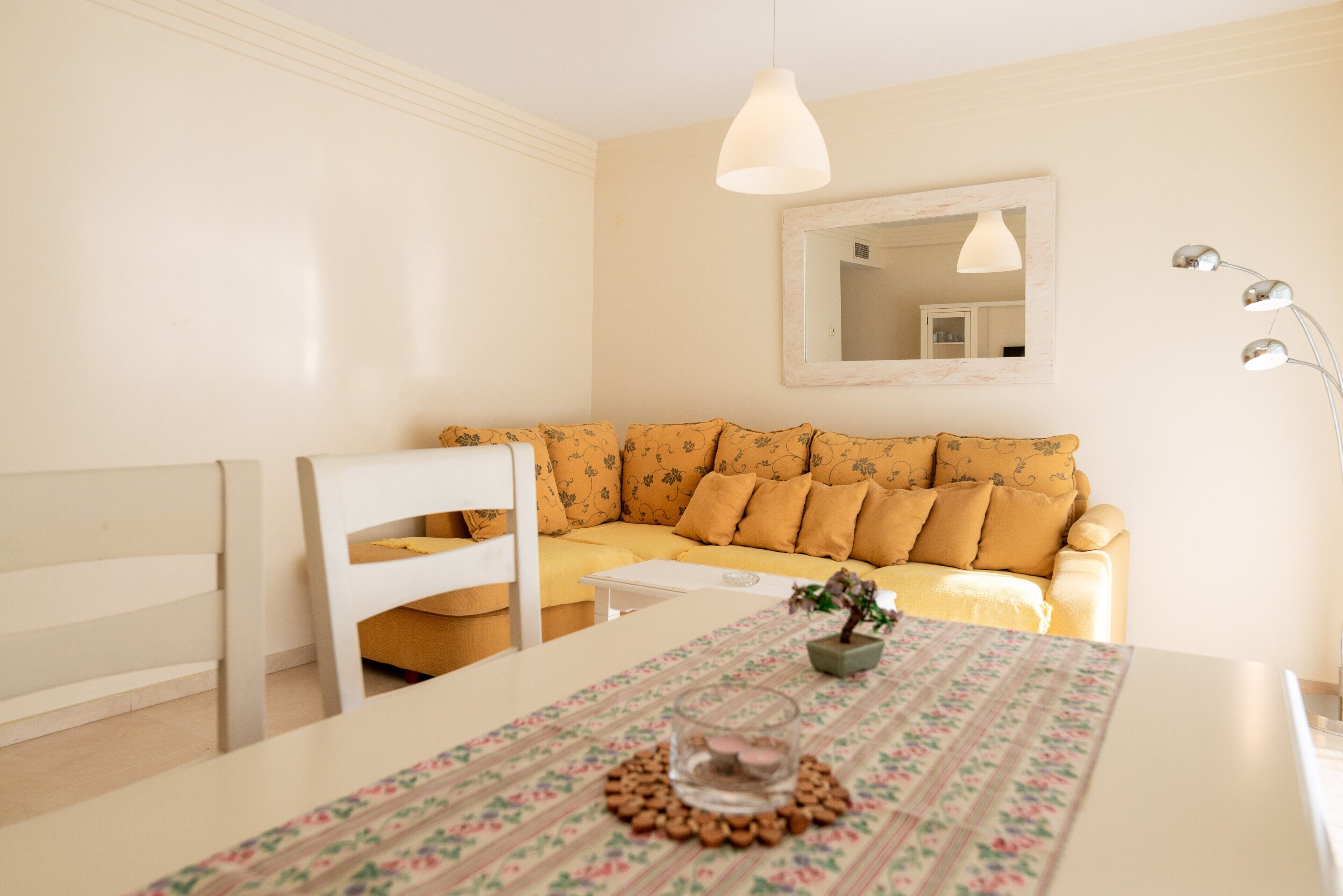 Ein Zimmer mit einem weißen Sofa, einem kleinen Tisch mit einer blumigen Tischdecke, einem Spiegel an der Wand und einer Pendelleuchte.
