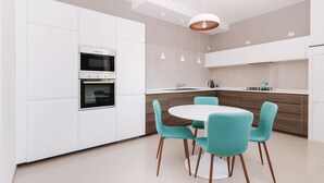 A modern kitchen with white cabinets, two ovens, a dining table with teal chairs, and pendant lighting. - Alchemy Apartment (Conversano)