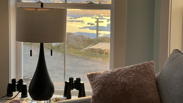 Habitació amb finestra gran que ofereix vista a un paisatge costaner, amb una làmpada, uns prismàtics i un coixí suau.