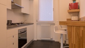 Una cocina moderna con armarios blancos, piso de baldosas negras, horno empotrado, estufa, ventana con persianas y encimera de madera con un microondas rojo y un tostador blanco.