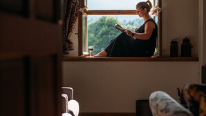 Une pièce où l’on voit une personne assise près d’une fenêtre, un canapé et une table avec une bougie.