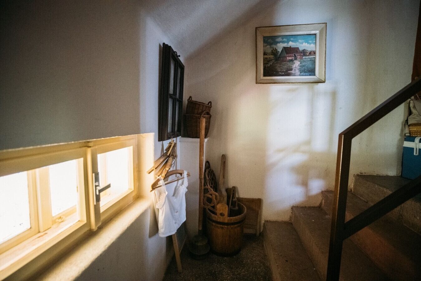 Uma escadaria estreita com corrimão de madeira, uma janela à esquerda e um quadro na parede.