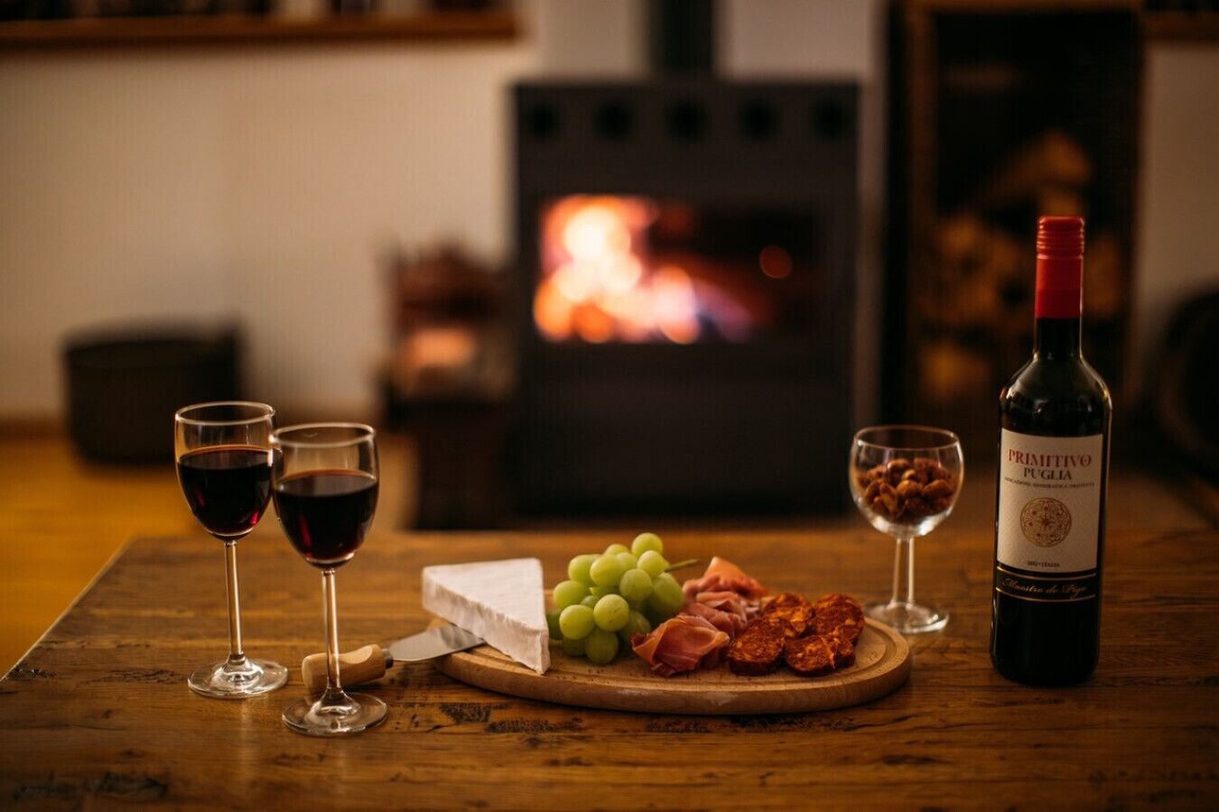 A wooden table with two wine glasses, a bottle of wine, a cheese wheel, grapes, and cured meats.
