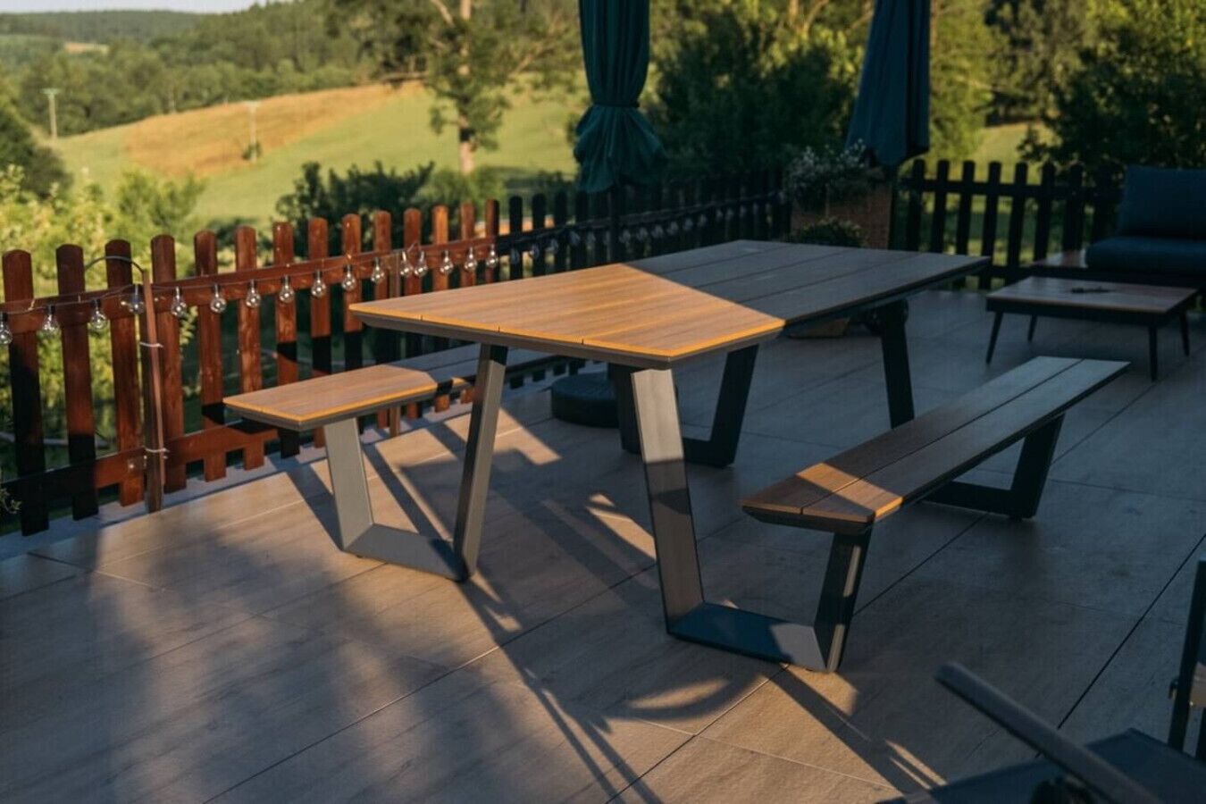An outdoor patio with a wooden table and benches, surrounded by a wooden fence and greenery.