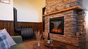 Un salon chaleureux avec une cheminée en pierre, un canapé vert, une petite table ronde ornée d’un vase et d’un verre à vin, et un mur en briques avec un tuyau noir.