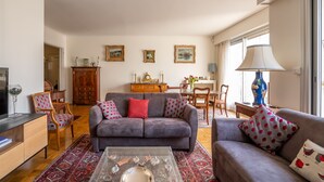 A living room with a grey sofa, red and white patterned cushions, a glass coffee table, and a wooden cabinet. There is a dining area with a table and chairs in the background.