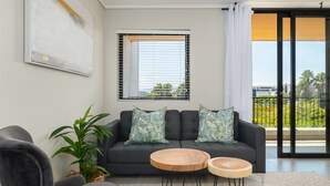 A modern living room with a grey sofa, two wooden side tables, a potted plant, and a large window with blinds. - Modern 1-Bedroom With Wetland Views | Netflix (Cape Town)