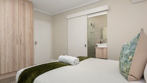 A hotel room with a bed, a wardrobe, a bathroom with a mirror, and a green carpet. - Modern 1-Bedroom With Wetland Views | Netflix (Cape Town)