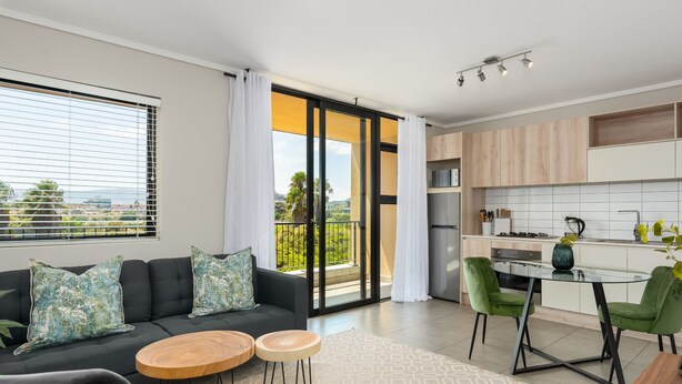 A modern living room with a sofa, two round wooden tables, and a dining area with green chairs. - Modern 1-Bedroom With Wetland Views | Netflix (Cape Town)