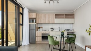 A modern kitchen with a dining area, featuring a glass table and green chairs, a refrigerator, and a view through large sliding doors. - Modern 1-Bedroom With Wetland Views | Netflix (Cape Town)
