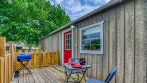 A covered outdoor patio with a wooden deck, a barbecue grill, a table with chairs, and a red door. - Kay's Cabin - Peaceful Acres (Rogersville)