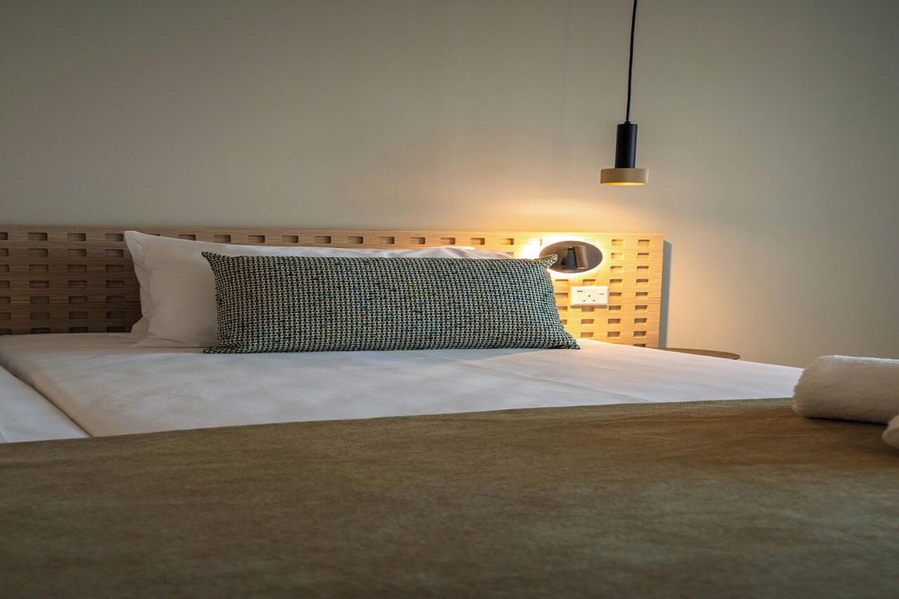 A neatly made bed with a patterned pillow, a round wall clock, and a hanging pendant light.