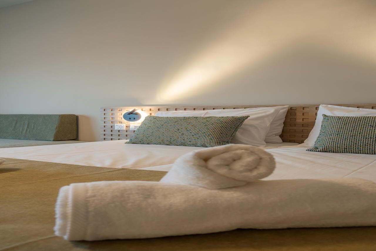 Una habitación de hotel con una cama, almohadas, una lámpara fijada en la pared y una toalla doblada con cuidado.