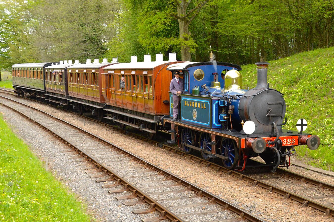 A vintage steam train with a blue and red locomotive, numbered 323, traveling on a single-track railway surrounded by greenery.