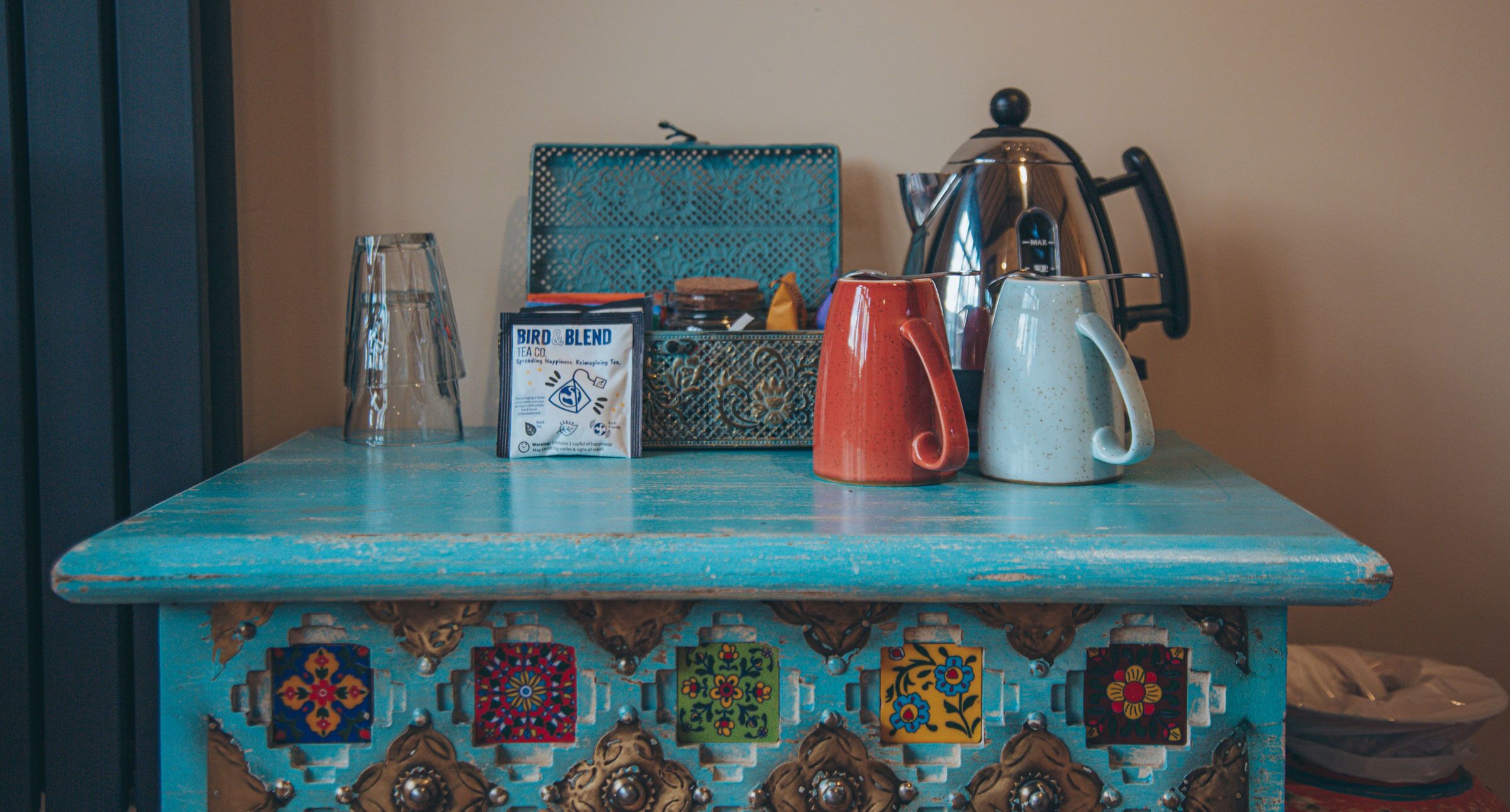 Uma mesa com superfície azul e azulejos decorativos, um copo, uma embalagem de chá, uma caneca vermelha, uma caneca branca e uma cafeteira.