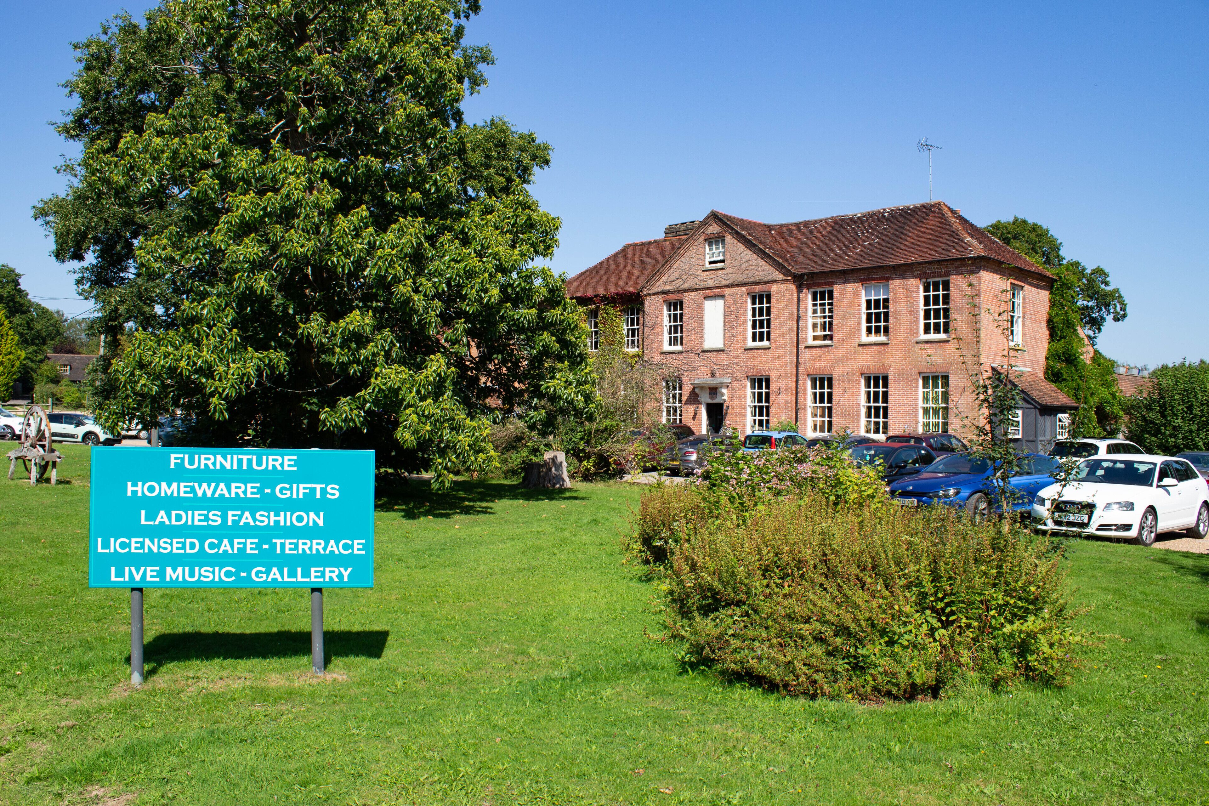 A two-story brick building with a traditional pitched roof, surrounded by lush greenery and parked cars.