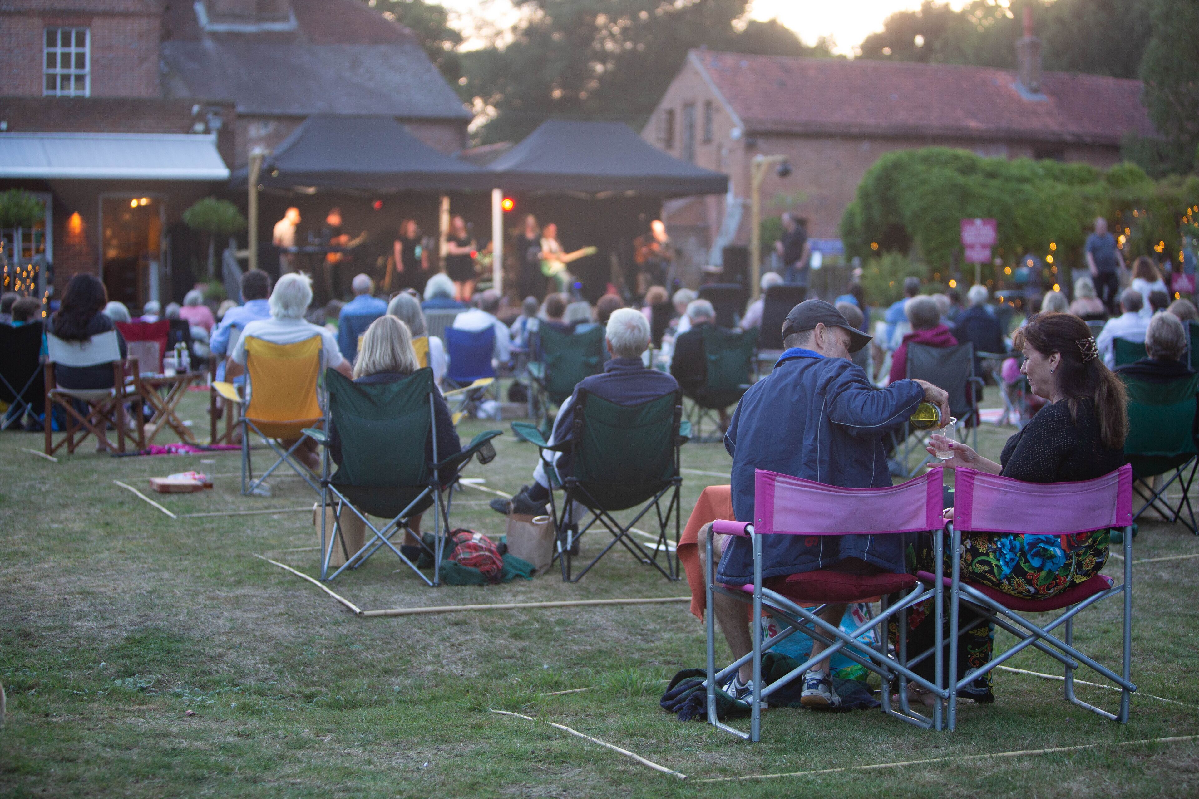 An outdoor gathering with people seated on folding chairs, a stage with musicians, and buildings in the background.