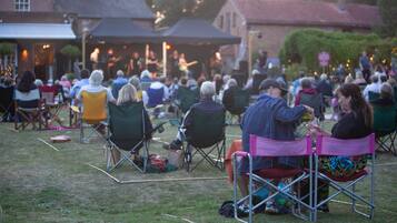 An outdoor gathering with people seated on folding chairs, a stage with musicians, and buildings in the background.
