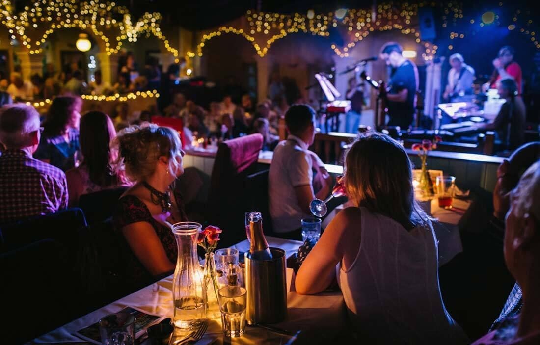 A crowded indoor venue with tables set for dining, a stage with musicians, and string lights overhead.