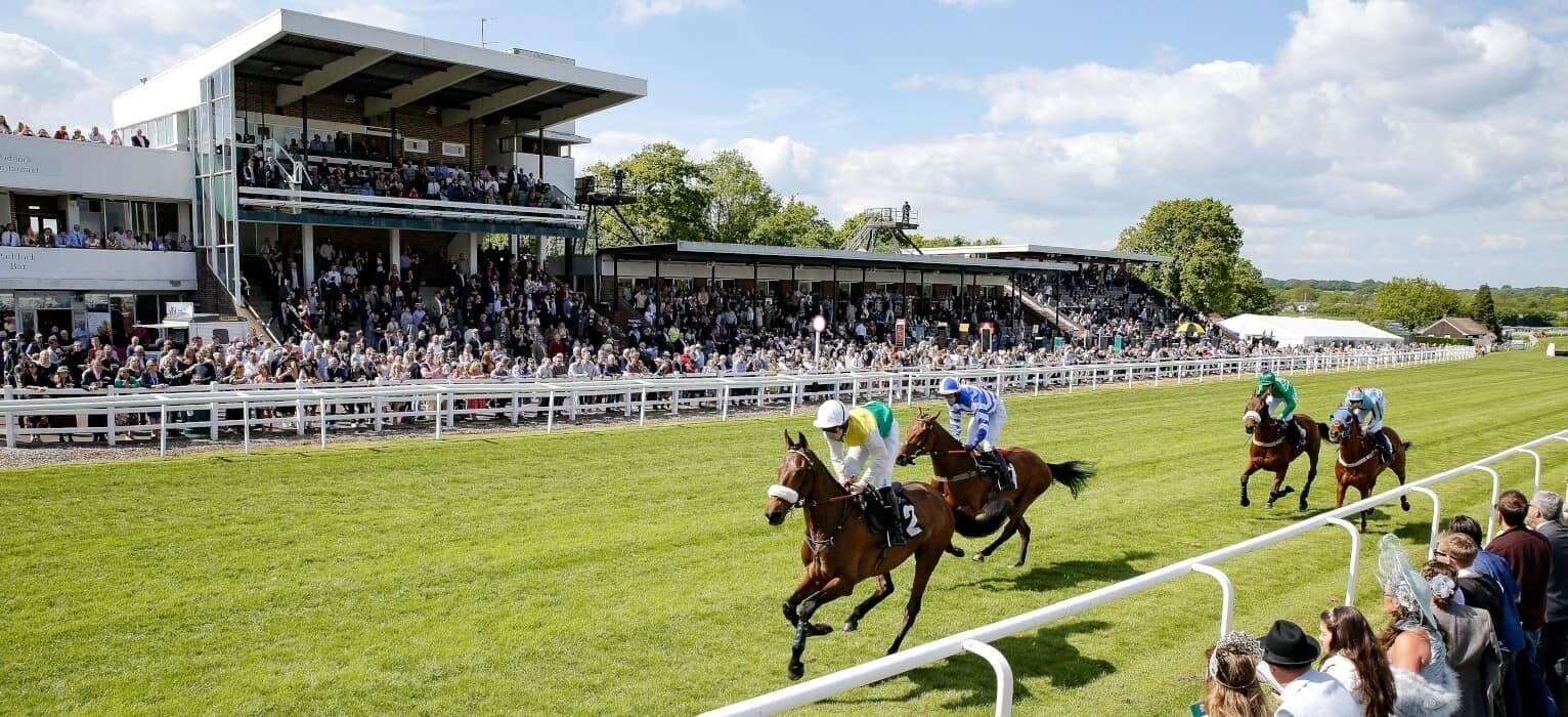 A horse race with jockeys on horses, spectators in the stands, and a large building with a covered area.