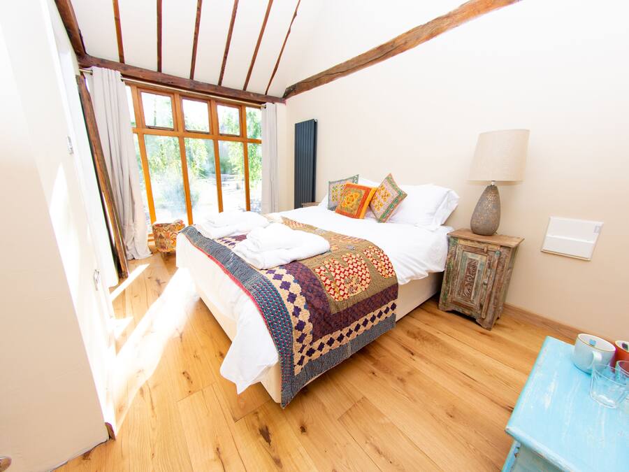 Quarto com piso de madeira, cama com manta estampada, mesa de cabeceira com abajur e vista para o verde lá fora.