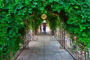 An arched pathway surrounded by lush green foliage, leading to a central area with string lights and a decorative element. - Trading Boundaries (Uckfield)