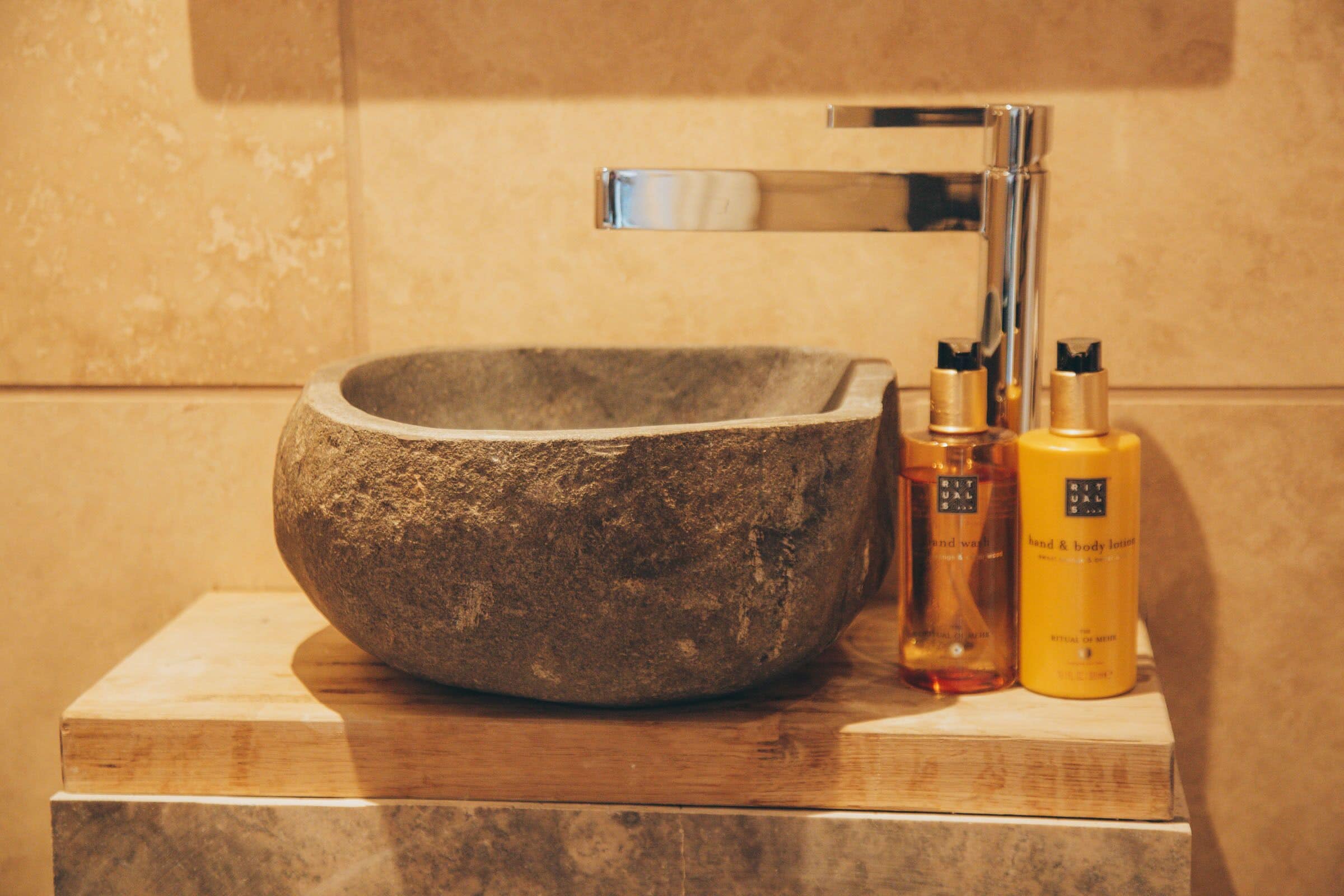 Un lavabo de piedra con grifo cromado, dos botellas de crema Ritual para manos y cuerpo, y una plataforma de madera.