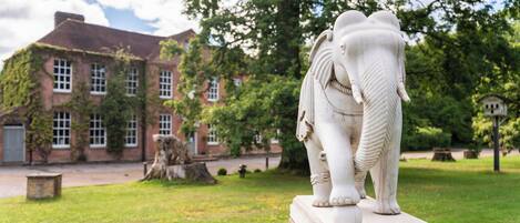 Una estatua blanca de elefante sobre un pedestal en una zona cubierta de césped, con un edificio al fondo.