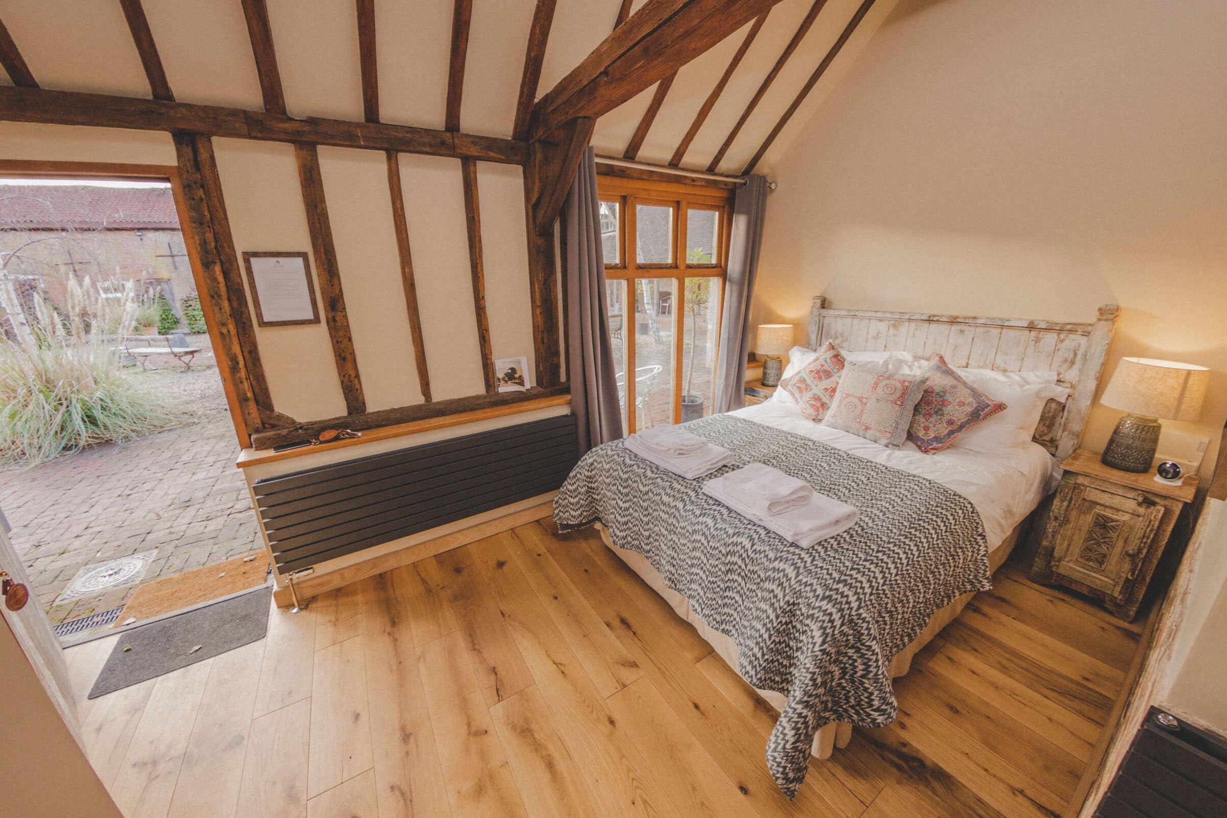 A bedroom with a wooden floor, a bed with patterned bedding, a wooden headboard, and a window with curtains.