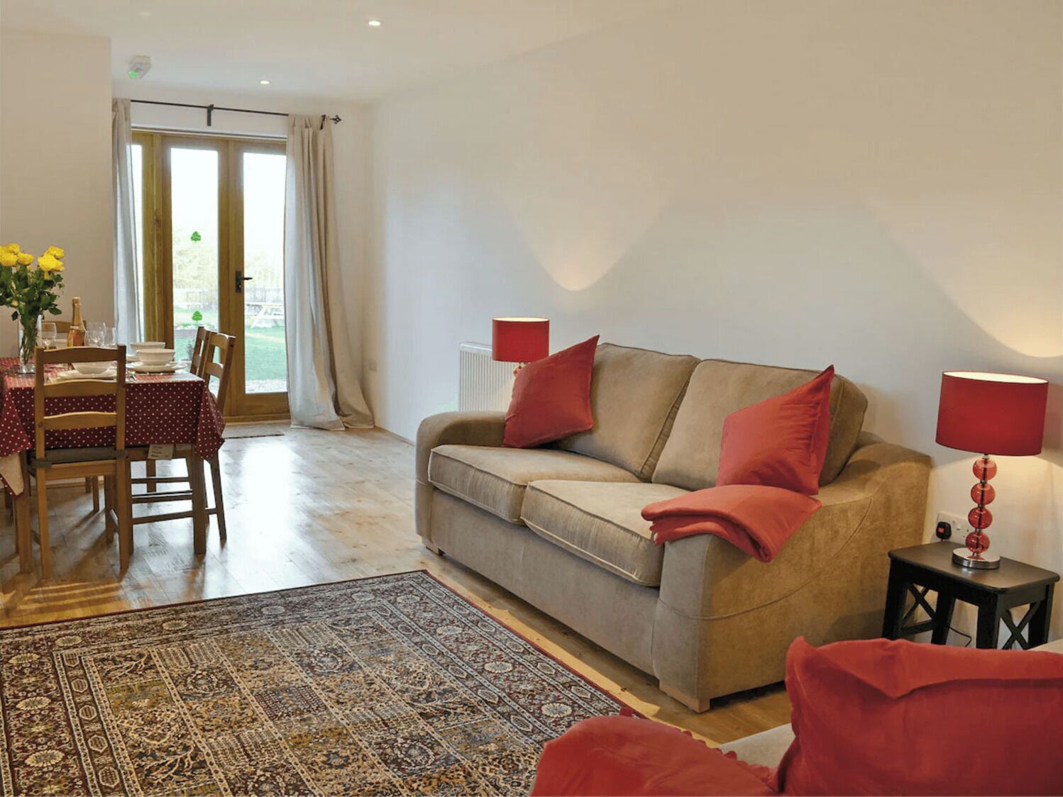 A living room with a beige sofa, red cushions, a patterned rug, a dining table with chairs, and a view through a glass door.