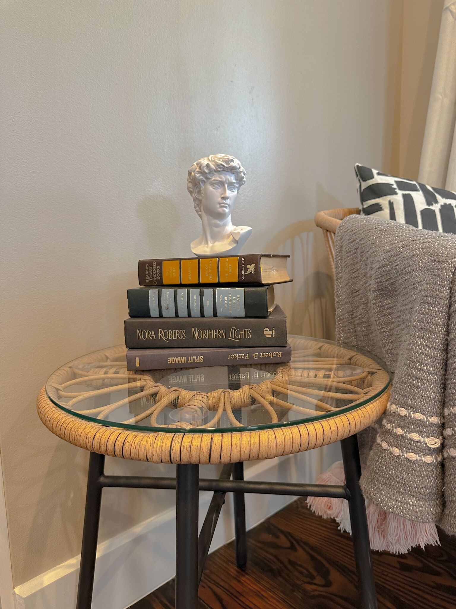 Ein Stapel Bücher auf einem Glastisch mit einem Flechtfuß, eine Büstenstatue und ein gemustertes Kissen auf einem Stuhl.