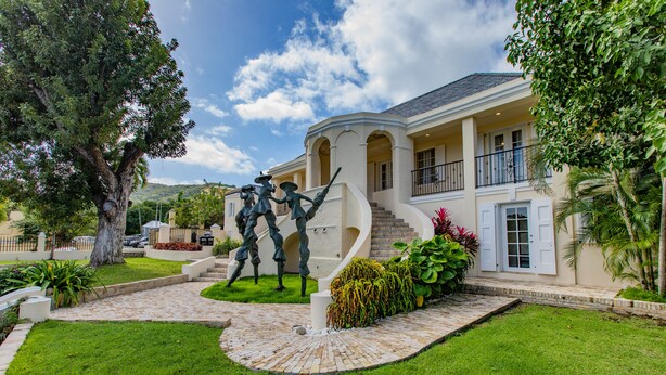 Um jardim de esculturas com uma casa, uma escadaria imponente, gramado bem cuidado e céu claro.