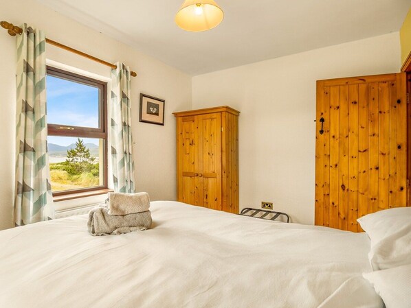 A bedroom with a bed, a window with curtains, a wooden wardrobe, and a wall-mounted light fixture. - Glynsk Pier Cottage (Cashel)