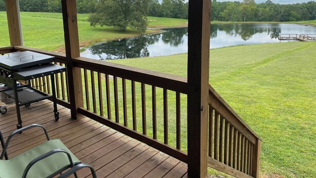 A covered outdoor area with a wooden deck, a grill, and a view of a lake and trees. - Cabin with private pond. A peaceful escape (Deer Lodge)