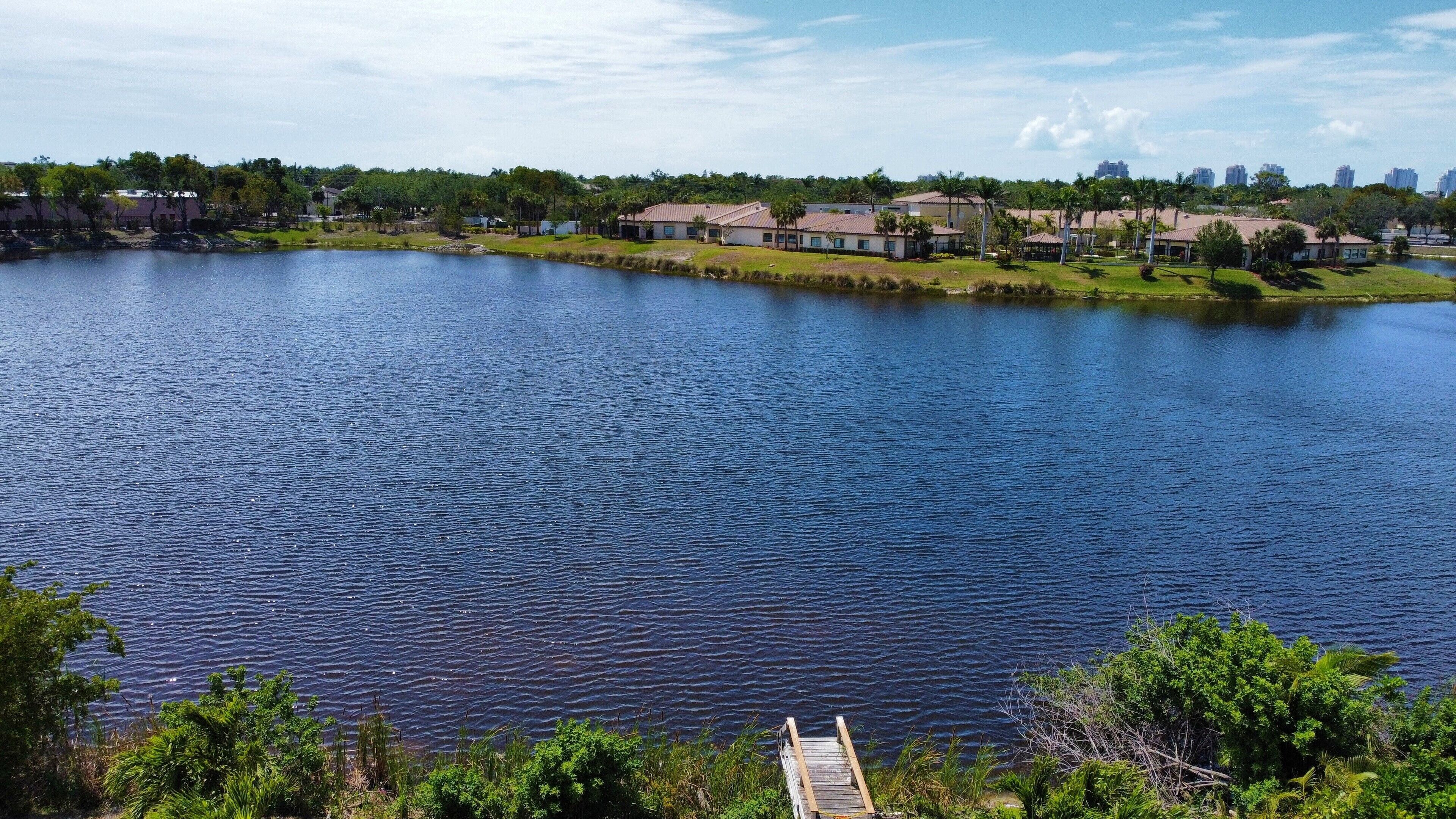 Un lago con un muelle, rodeado de vegetación y edificios residenciales al fondo.