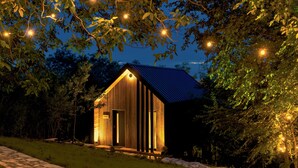 A wooden cabin with a sloped roof, surrounded by trees and illuminated by string lights.