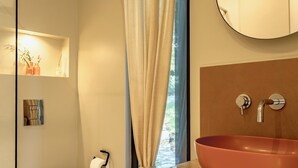 A modern bathroom with a white toilet, a beige shower curtain, a round mirror, and a wooden vanity with a red basin.
