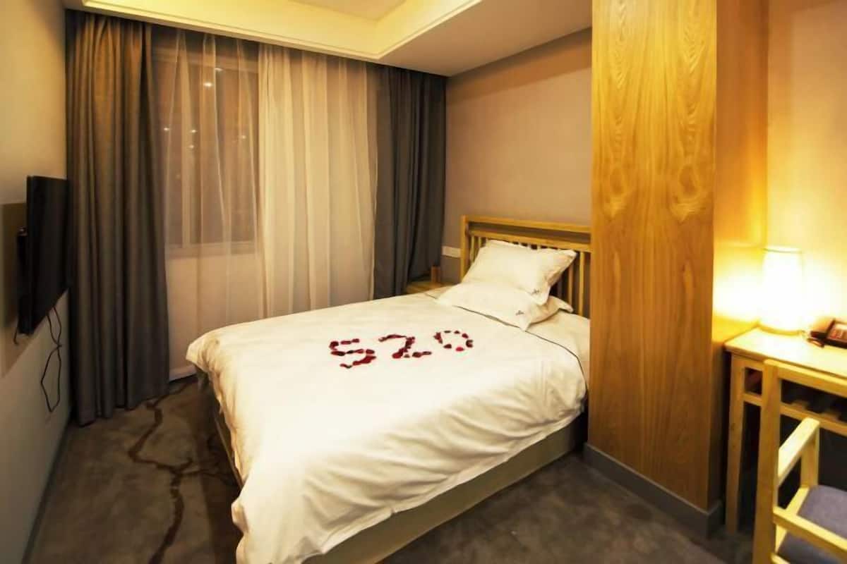 Ein Hotelzimmer mit Bett, Vorhängen, einem Holz-Kopfteil, einem Nachttisch mit Lampe und einem an der Wand befestigten Fernseher.