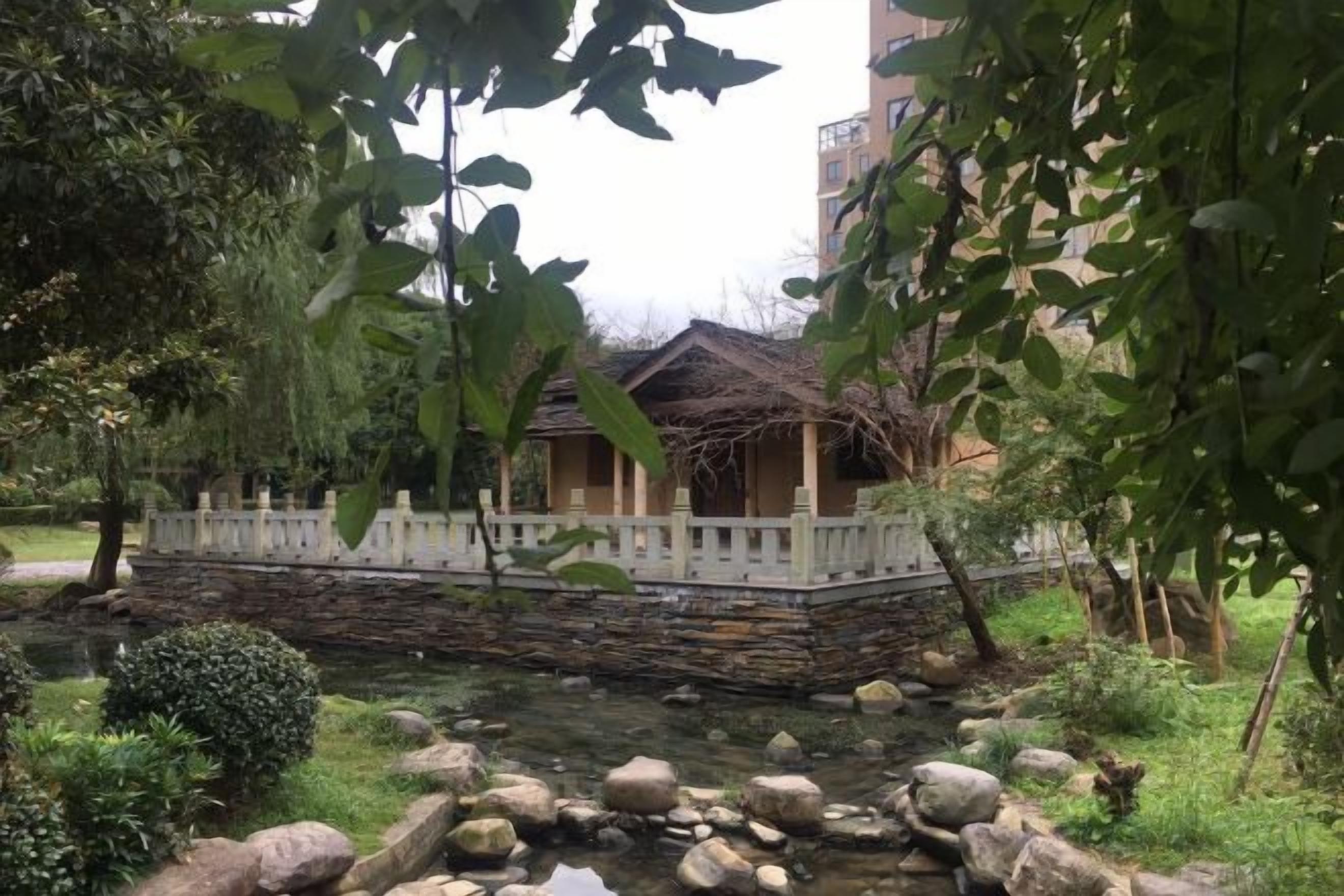Un edificio de estilo tradicional con techo de paja, rodeado por un muro de piedra y una valla blanca, situado junto a un pequeño arroyo con rocas y vegetación.