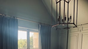 Een slaapkamer met een groot bed, een stoel, een kroonluchter en uitzicht door een raam met gordijnen.
