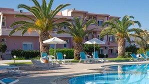A pool area with lounge chairs and umbrellas, palm trees, and a pink building in the background. - SEA SIDE (Chania)