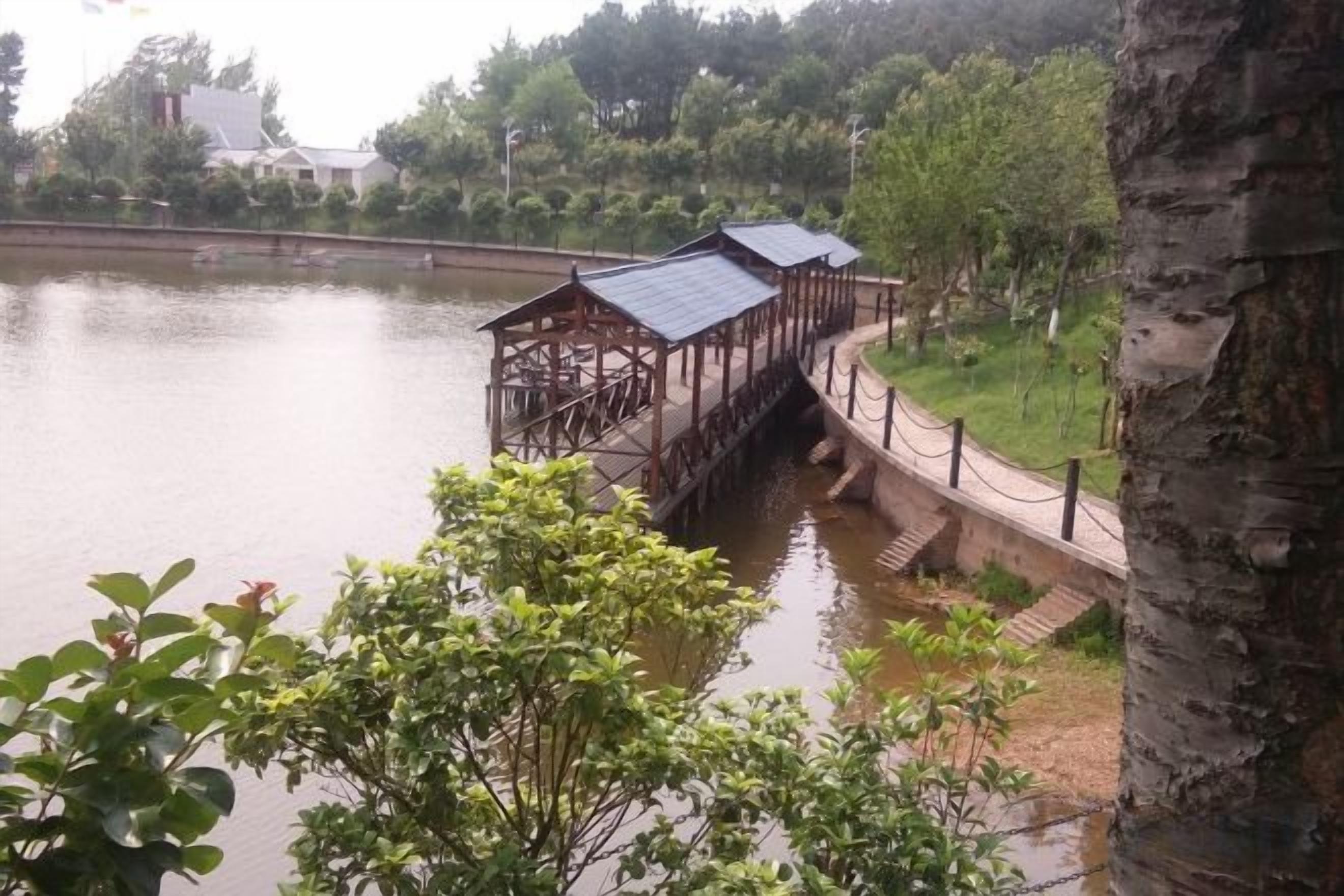 A wooden bridge over a calm body of water, surrounded by greenery and a building in the background.