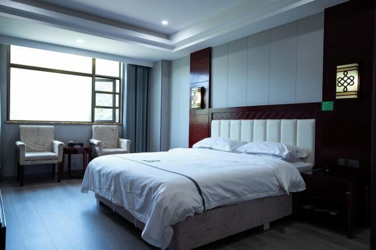 A hotel room with a large bed, two armchairs, a small table, and a window providing natural light.