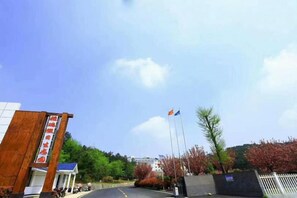 A building with a sign, a flagpole with flags, and a parking area with trees and a fence. - Yarui Holiday Garden Hotel (Guangshui)