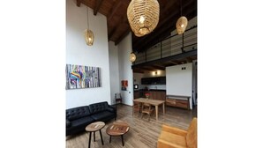 Un'ampia zona giorno con un divano nero, due tavolini in legno e una poltrona gialla. Un tavolo da pranzo in legno è apparecchiato con un vaso di fiori. La stanza presenta travi a vista in legno e un grande quadro astratto appeso alla parete.