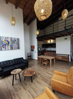 Een ruime woonkamer met een zwarte bank, twee houten bijzettafels en een gele fauteuil. Een houten eettafel is gedekt met een vaas bloemen. De kamer heeft zichtbare houten balken en een groot abstract schilderij aan de muur.