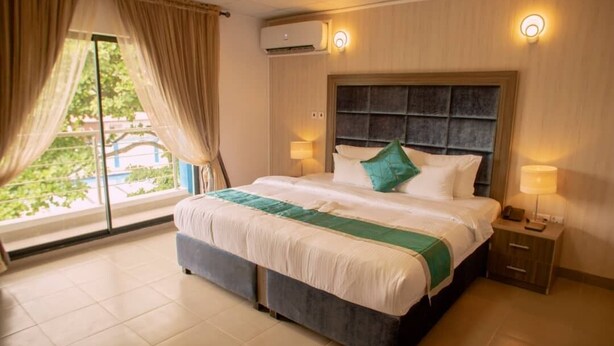 A hotel room with a large bed, two bedside lamps, an air conditioning unit, and a view through the window. - Riviera Suites (Lagos)