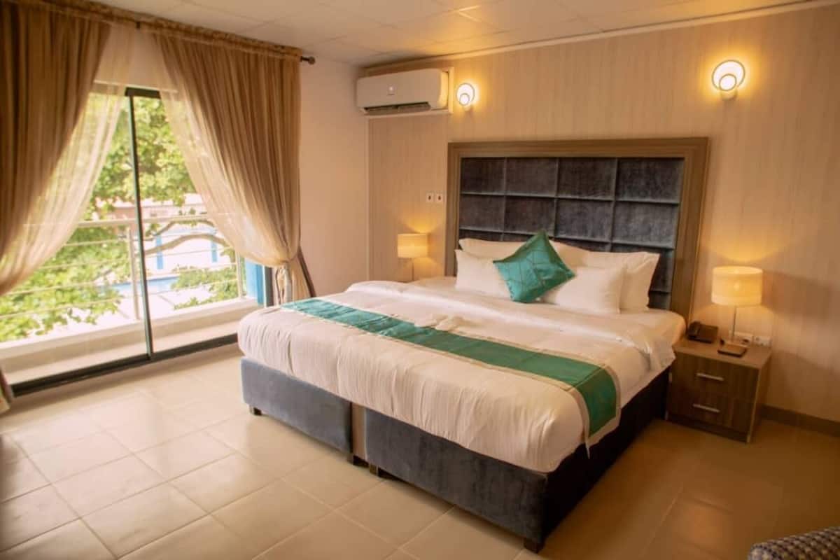A hotel room with a large bed, two bedside lamps, an air conditioning unit, and a view through the window.