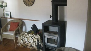 Ein gemütliches Wohnzimmer mit Kamin, einem Korbstuhl, einem kleinen Tisch mit Vase und Bilderrahmen sowie einer Uhr an der Wand.